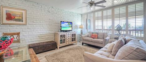 A living room with a white brick wall, a ceiling fan, a television, and a large window with blinds.