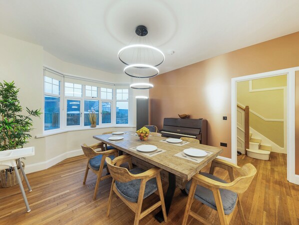 A dining area with a wooden table, six chairs, a piano, and a large window.