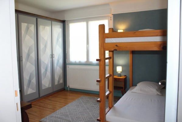 A bedroom with a bunk bed, a wardrobe with frosted glass doors, a small bedside table with a lamp, and a window providing natural light.