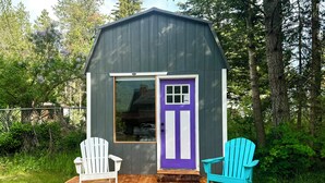 Un cobertizo de madera pequeño con una puerta morada y ventanas, rodeado por una terraza de madera con dos sillas Adirondack.