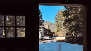 Vista a través de una ventana que muestra una casa con patio nevado, una terraza de madera y una montaña al fondo.