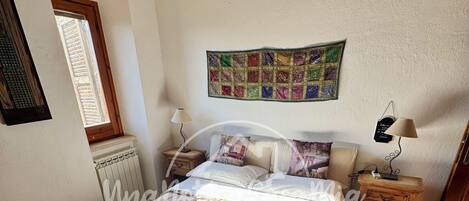 Ein Schlafzimmer mit einem großen Bett, einem Fenster, einer Heizung, einer Wandteppich und einem Nachttisch mit einer Lampe.