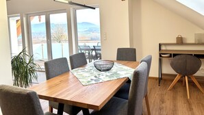 A dining area with a wooden table, grey upholstered chairs, and a view of a lake through large windows. - "Seeblick" guest house - Bright penthouse apartment on the outskirts of Salzburg (Seekirchen am Wallersee)