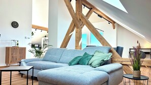 A modern living room with a grey sectional sofa, a small black table, and a round wireframe side table. A wooden ceiling structure and a skylight are visible. - "Seeblick" guest house - Bright penthouse apartment on the outskirts of Salzburg (Seekirchen am Wallersee)