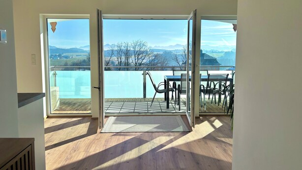 A room with a wooden floor, a dining table with chairs, and a view of a lake and mountains through large glass doors. - "Seeblick" guest house - Bright penthouse apartment on the outskirts of Salzburg (Seekirchen am Wallersee)
