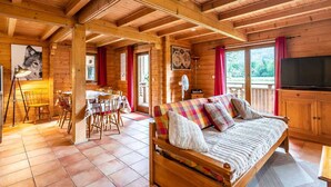 L’intérieur chaleureux d’un chalet avec un plafond en bois, un coin repas comprenant une table et des chaises, un canapé agrémenté de coussins colorés et un téléviseur.