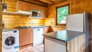 Une cuisine compacte aux murs en bois, équipée d’un four à micro-ondes, d’un réfrigérateur, d’un lave-linge et d’une fenêtre donnant sur un paysage verdoyant.
