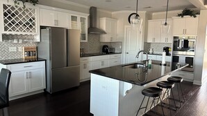 A modern kitchen with white cabinetry, a stainless steel refrigerator, a double oven, and a large island with bar stools. - Master's Tournament Rental House (Grovetown)