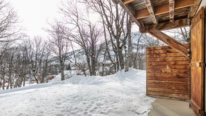 Une cabane en bois, le sol recouvert de neige, avec en arrière-plan des arbres dénudés et des montagnes.