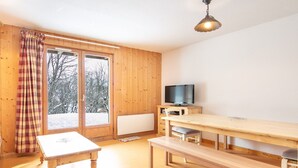Une pièce chaleureuse aux murs et au plancher en bois, avec une grande fenêtre donnant sur les arbres, un téléviseur et une longue table en bois.