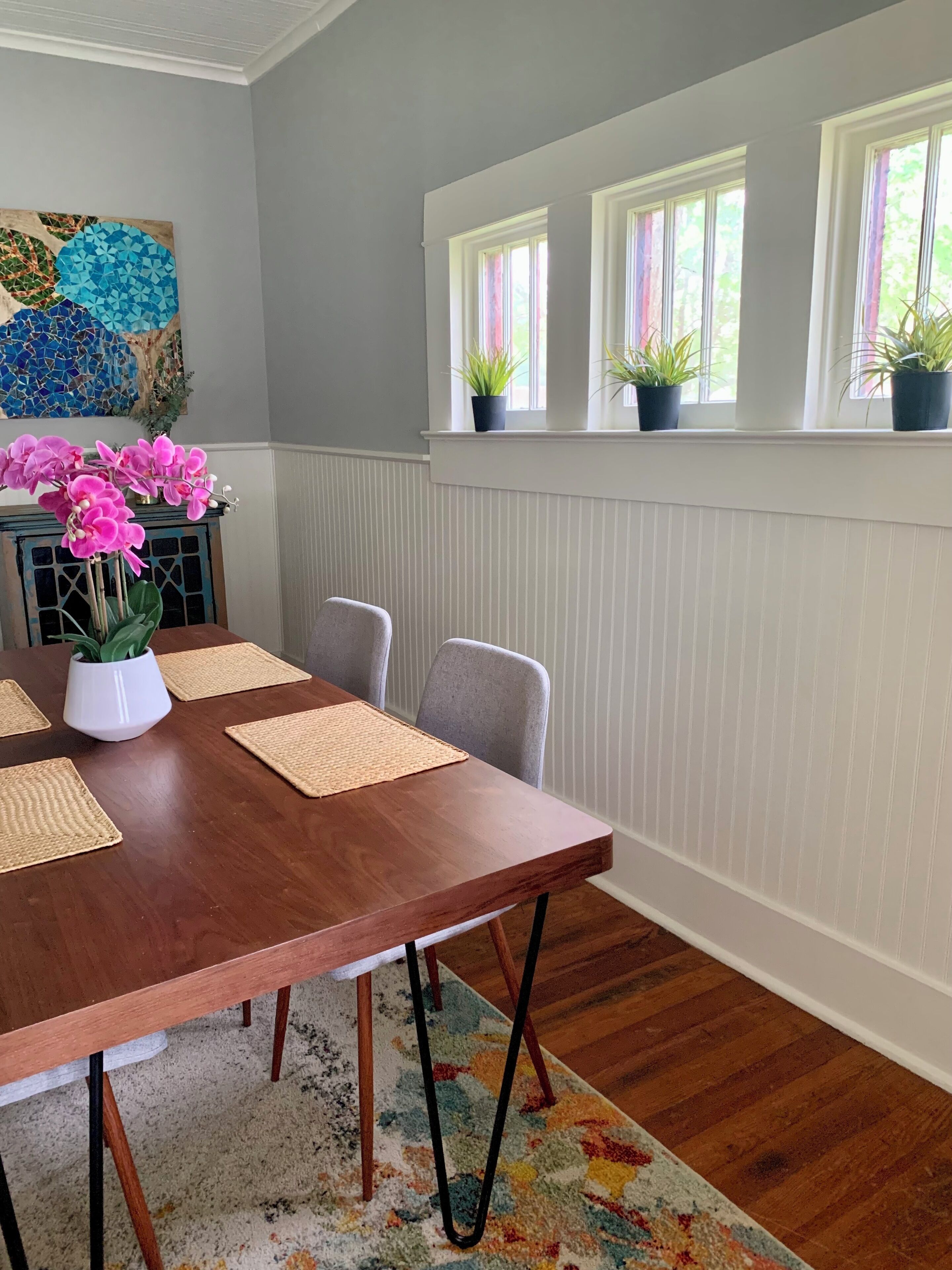 A dining room with a wooden table, four chairs, a vase with pink flowers, and a colorful rug.