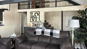 Ein geräumiges Wohnzimmer mit einer braunen Ledersofa, einer karierten Tagesdecke und einem an der Wand befestigten 'LOVE'-Schild. Eine Treppe führt in ein Obergeschoss, und eine Pflanze ist in der Ecke zu sehen.