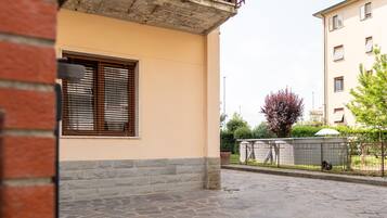 A building with a stone-paved courtyard, a window with blinds, and a fenced area with greenery.