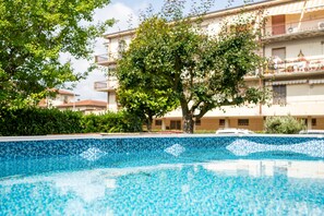 A swimming pool with a tiled edge, surrounded by greenery and a multi-story building in the background.