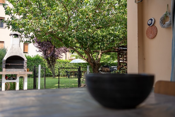 A patio with a stone barbecue grill, a table with a black bowl, and a tree providing shade.