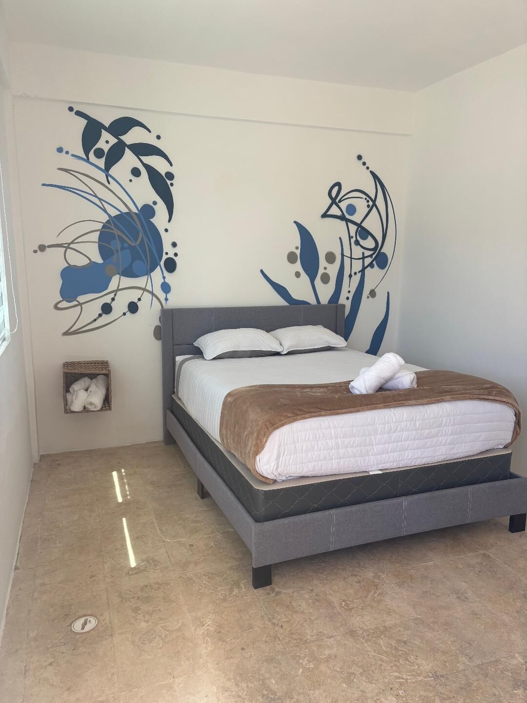 Un dormitorio con una cama, una pared decorada con motivos florales abstractos, una cesta pequeña con toallas y un suelo de baldosas.