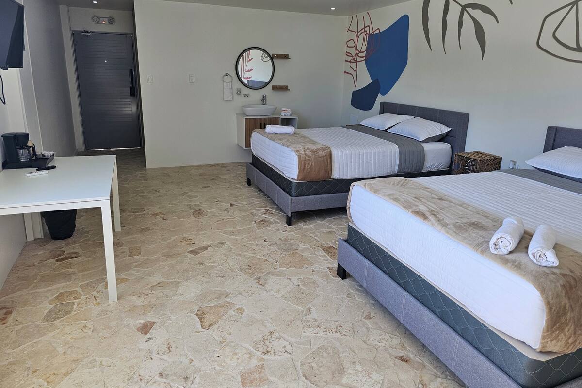 A hotel room with two beds, a desk, a bathroom, and a stone-tiled floor.