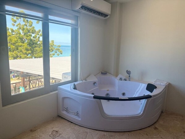 A modern bathroom with a large bathtub, a window with a view of the beach, and a ceiling air conditioner. - Buye Apartments (Cabo Rojo)