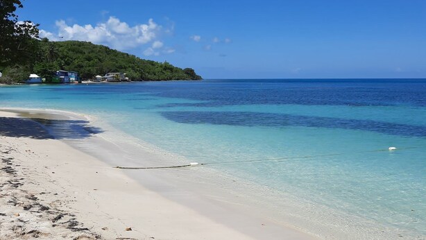 A beach with clear turquoise water, a sandy shore, and a small hill with buildings in the background. - Buye Apartments (Cabo Rojo)