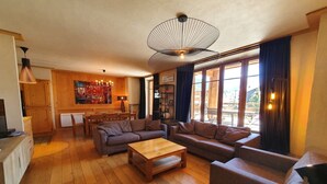 Un salon chaleureux avec un plancher en bois, une grande table basse en bois et deux canapés. On y trouve également un coin repas avec une table et des chaises, ainsi qu’une cuisine équipée d’un réfrigérateur et de placards.