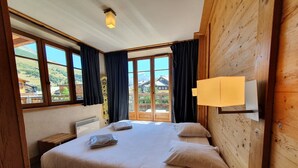 A wooden-paneled room with a large bed, a nightstand, a lamp, and a view of a mountain and buildings through the windows. - Alpine Comfort for 8 guests (Les Deux Alpes)