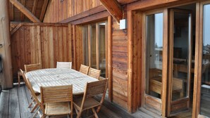 Una terraza de madera con una mesa y sillas, grandes puertas correderas de vidrio y una estructura de techo de madera.