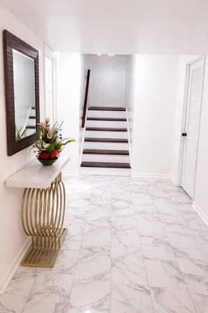 Un pasillo con piso de mármol, una consola blanca con un florero y un espejo grande, además de una escalera.