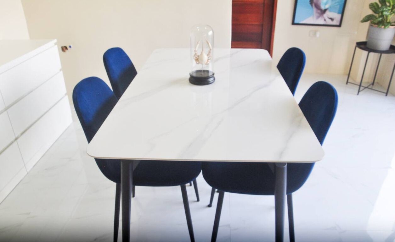 Uma mesa de jantar com seis cadeiras azuis, um centro de mesa em cúpula de vidro transparente e superfície de mármore branco.