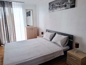 A neatly made bed with a patterned bedspread, a nightstand with a lamp, a wooden wardrobe, and a cityscape painting on the wall. - Comfortable Apartment With sea View (Bar)