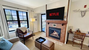 Una sala de estar moderna con chimenea, una pantalla grande mostrando 'NETFLIX' y vistas al exterior a través de amplias ventanas.