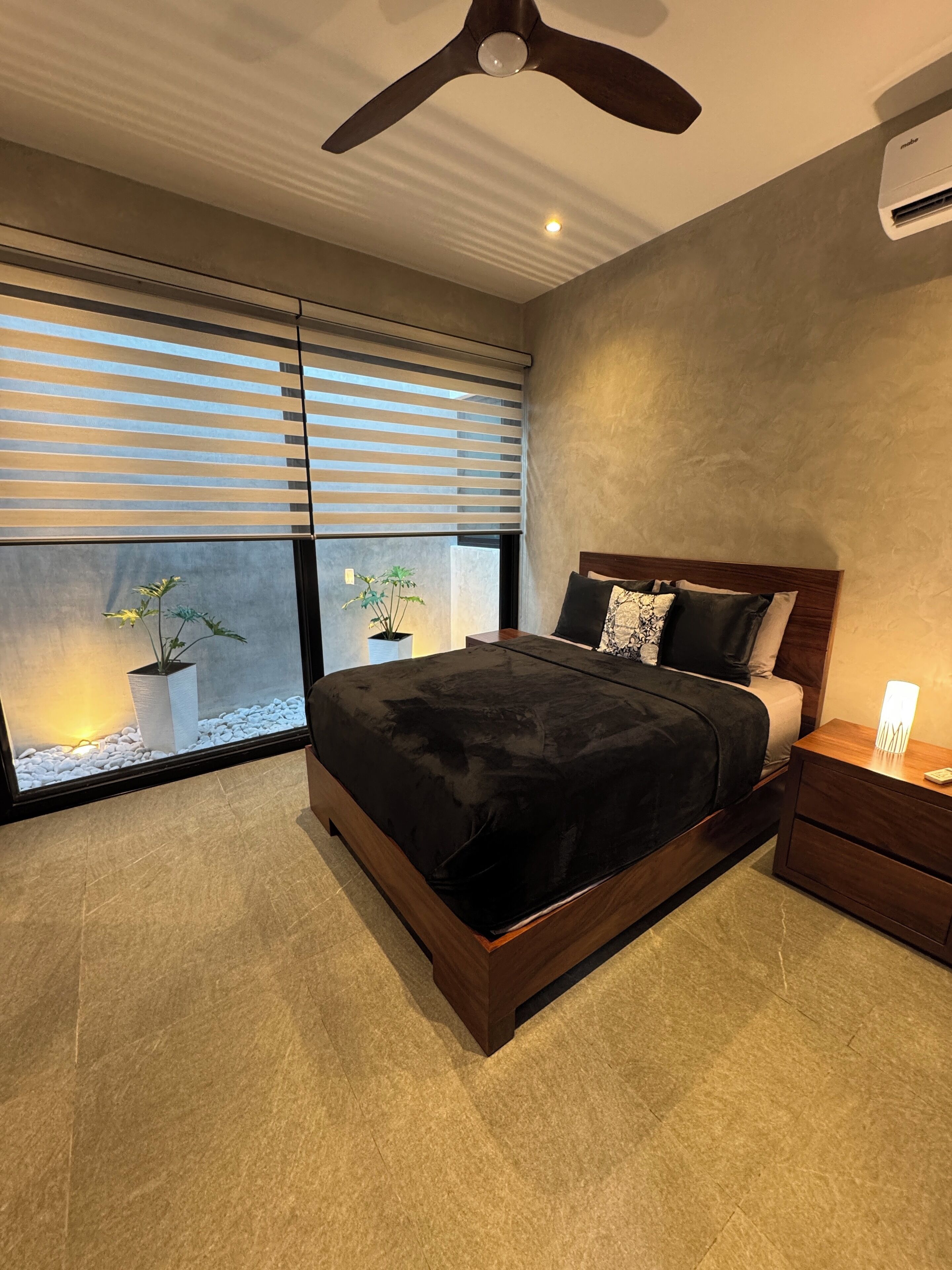Een moderne slaapkamer met een groot bed, een plafondventilator, een nachtkastje en een raam met jaloezieën.