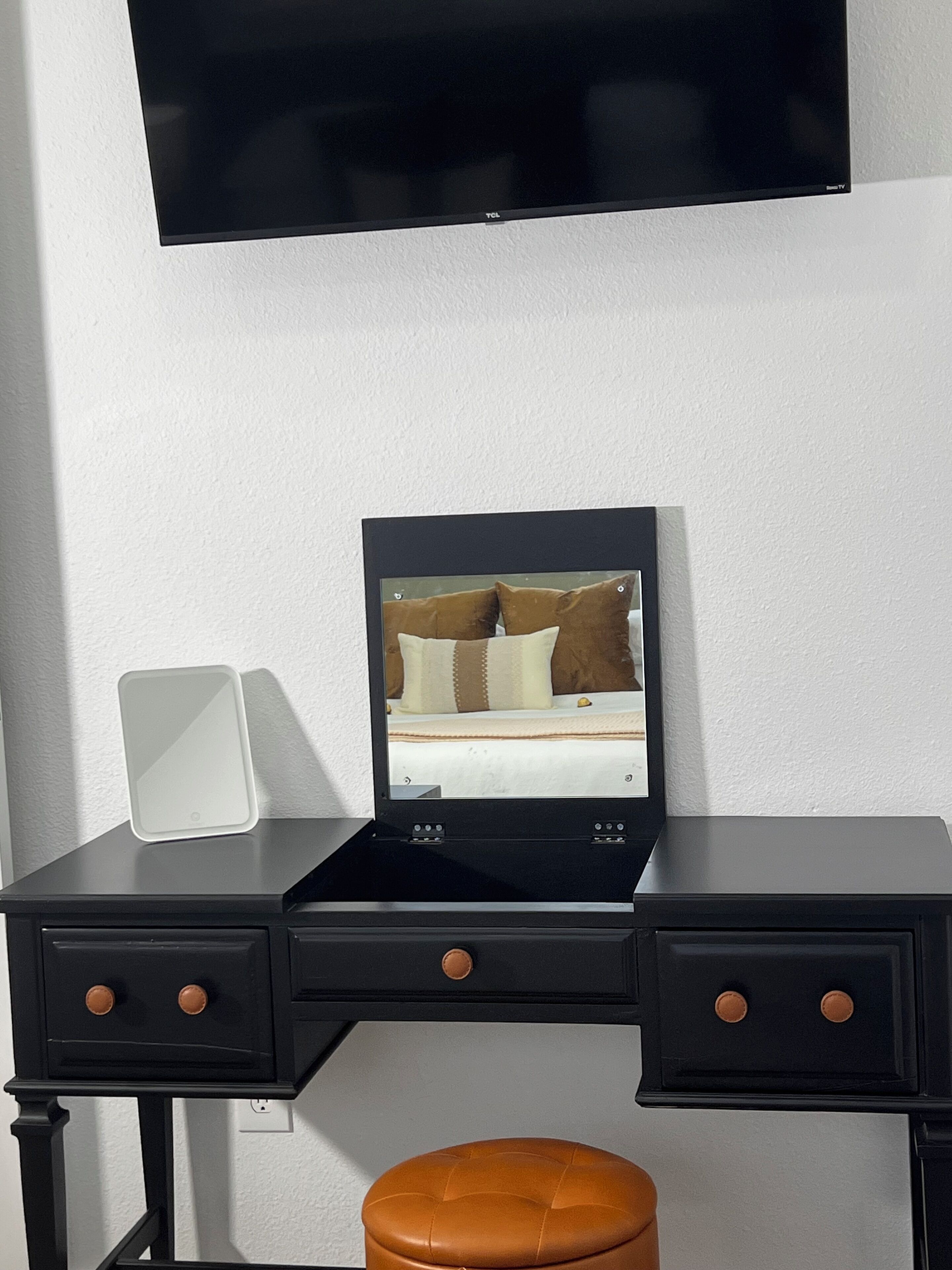 A hotel room with a black desk, a flat-screen TV mounted on the wall, a tablet on the desk, and a brown leather ottoman.
