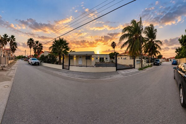 A street view at sunset with a gated building, palm trees, and parked cars.
