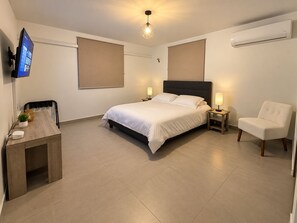 Un dormitorio moderno con cama, mesitas de noche, una silla, escritorio, televisor y aire acondicionado.
