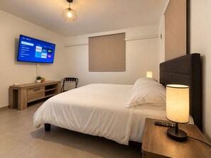 Une chambre d’hôtel comprenant un lit, un téléviseur fixé au mur, une table de chevet avec une lampe et un petit bureau avec une chaise.