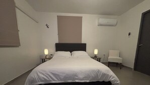 Cama bem arrumada em quarto de hotel, com cabeceira, duas mesas de cabeceira com abajures, uma cadeira e um aparelho de ar condicionado na parede.