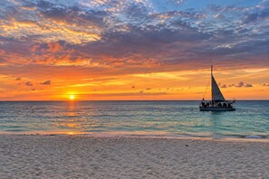 Escena de playa con un velero en aguas tranquilas durante la puesta de sol.