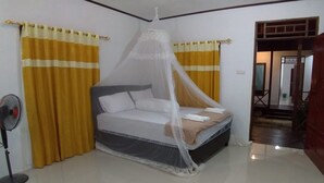 Un dormitorio con cama, cortinas amarillas, un ventilador de pie y una puerta que da a otra habitación.