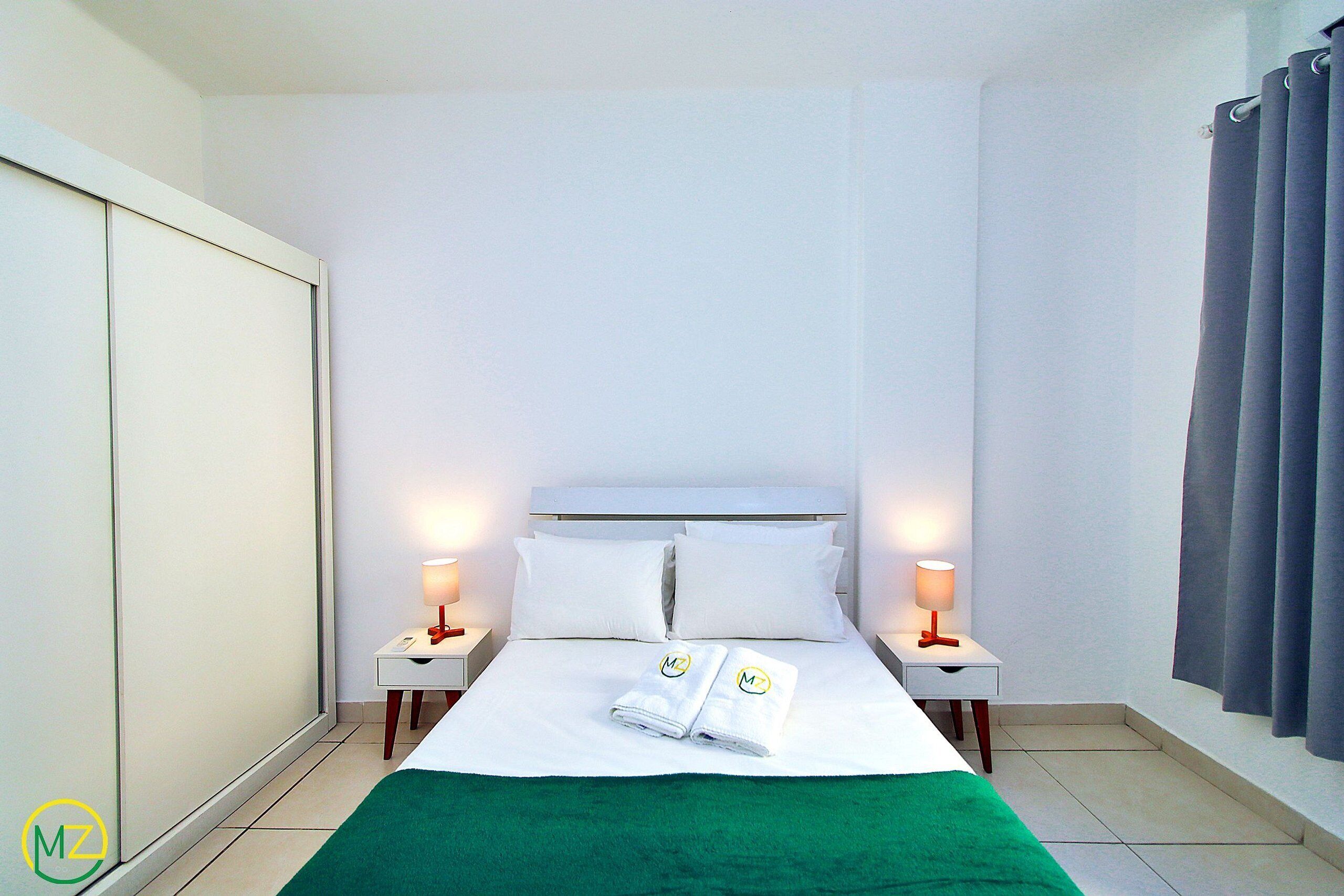 Quarto de hotel com uma cama, duas mesas de cabeceira com luminárias, um cobertor verde e um guarda-roupa.