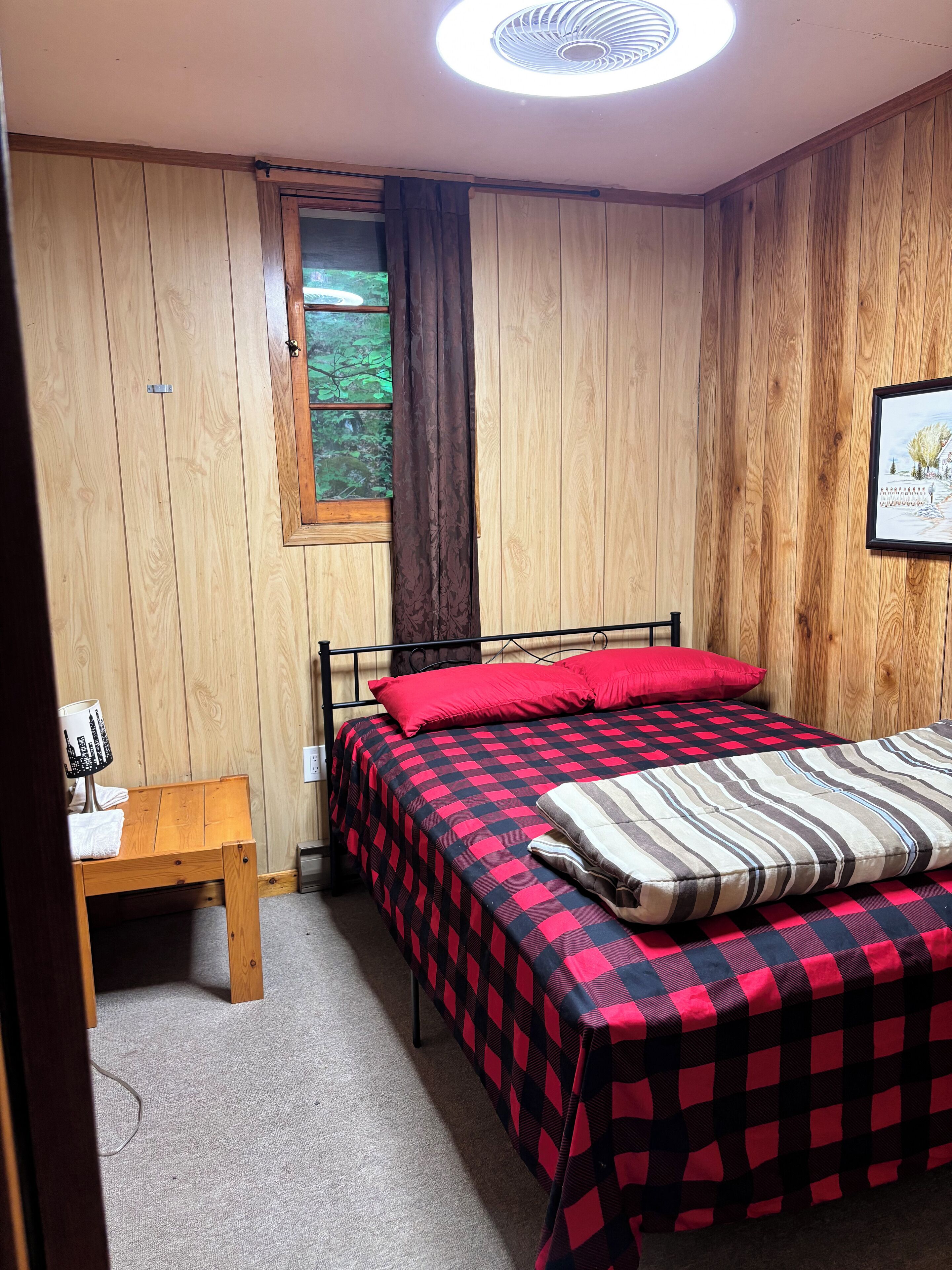 A room with wooden walls, a bed with a checkered bedspread, a window with curtains, a bedside table with a lamp, and a framed picture on the wall.