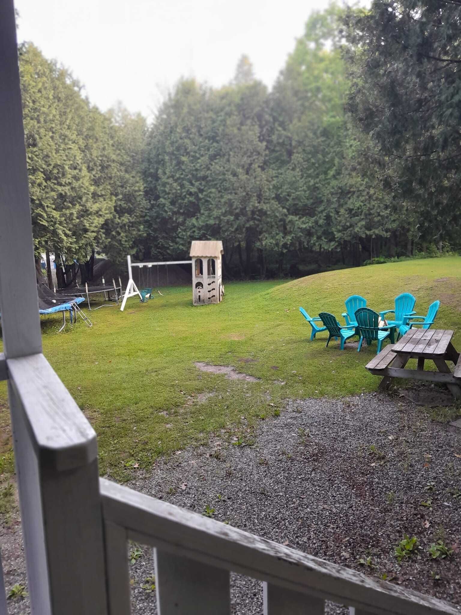 Un espace extérieur verdoyant, avec une petite structure en bois, qui pourrait être une cabane de jeux, ainsi que plusieurs chaises en plastique vertes et une table de pique-nique.