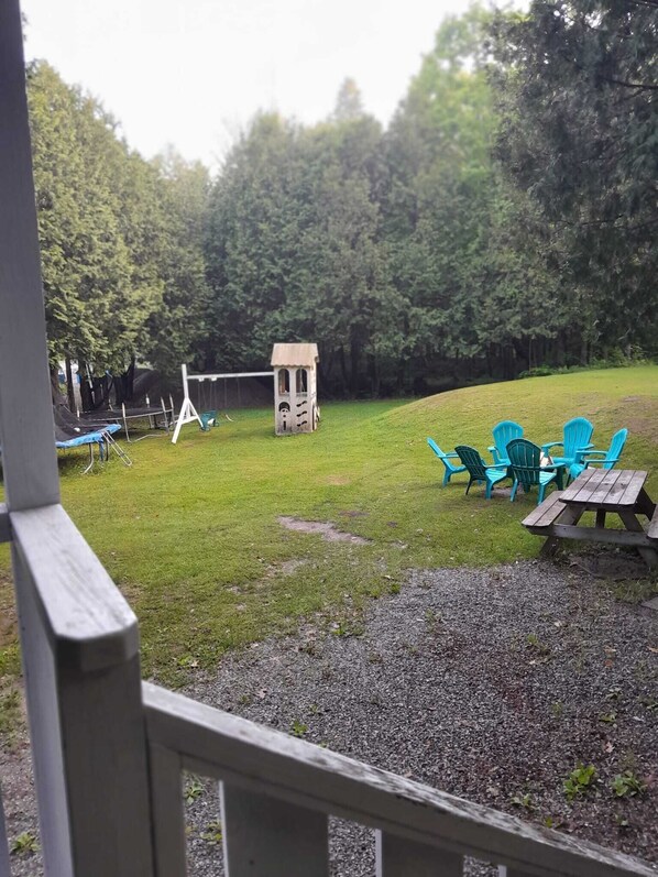 Un espace extérieur verdoyant, avec une petite structure en bois, qui pourrait être une cabane de jeux, ainsi que plusieurs chaises en plastique vertes et une table de pique-nique.