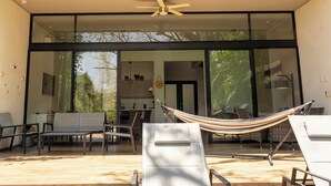 A modern outdoor seating area with a hammock, lounge chairs, and a dining area visible through large glass doors. - New House in Santa Fe Morelos (Xochitepec)
