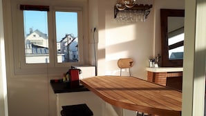 Une cuisine avec une table en bois, deux tabourets de bar, une fenêtre donnant sur des immeubles et un porte-bouteilles fixé au mur.