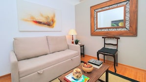 A living room with a beige sofa, a wooden coffee table with a bowl of fruit, a framed mirror, and a painting on the wall. - Nossa Senhora VII (Rio de Janeiro)
