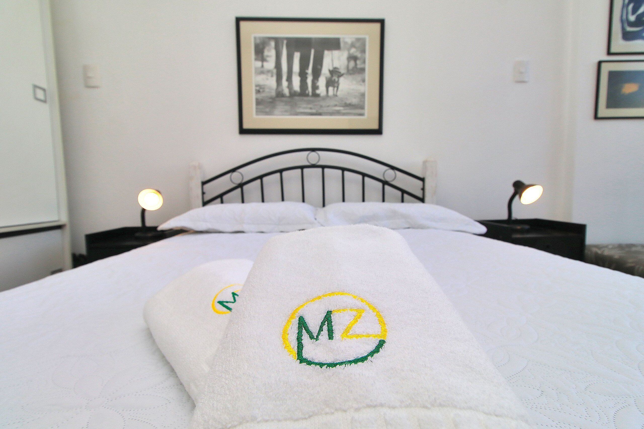A neatly made bed with a white duvet and two towels with a logo on them. There is a framed picture on the wall above the bed.