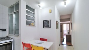 A small kitchen with a gas stove, a window with blinds, a white table with two chairs, and a framed picture on the wall. - Nossa Senhora VII (Rio de Janeiro)