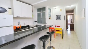 A modern kitchen with white cabinets, a black countertop, and a bar stool. There is a dining area with a yellow table and red chairs. - Nossa Senhora VII (Rio de Janeiro)