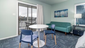 Una habitación de hotel con balcón, una mesa redonda con sillas, un sofá verde, una mesita de noche y un cuadro en la pared.