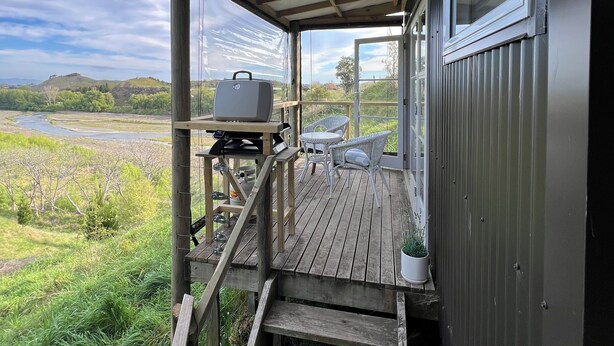 A wooden deck with a grill and chairs, overlooking a river and hills. - Quiet, private Cosy Cabin with en-suite. Access to bike paths, river and pub. (Puketapu)